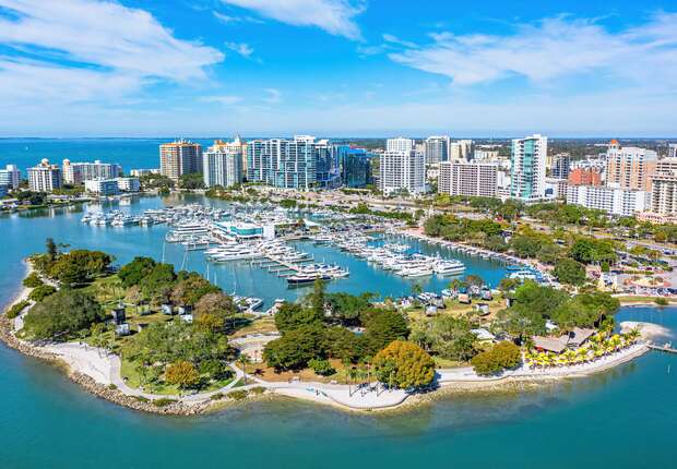 A marina filled with numerous boats is surrounded by lush green parks. In the background, a cityscape with tall buildings lines the waterfront under a clear blue sky.