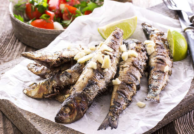 Grilled sardines are laid on parchment paper, garnished with chopped garlic and lime wedges. In the background, a bowl of fresh tomato and herb salad adds color to the wooden table setting.
