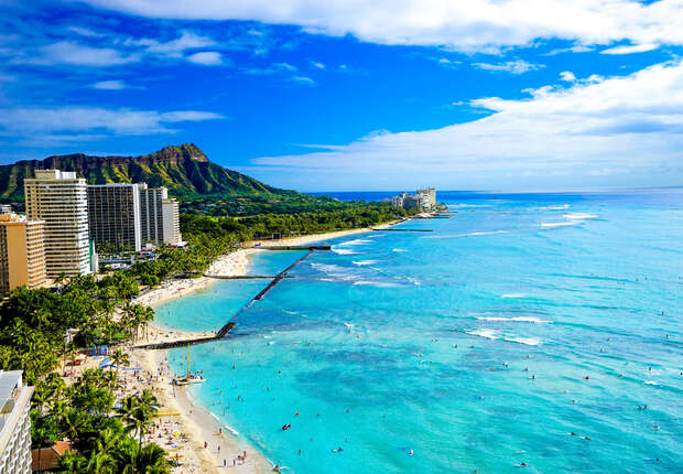 A sandy beach overlooks a turquoise ocean with surfers and swimmers. Skyscrapers line the shore, and a lush green mountain rises in the background under a bright blue sky.