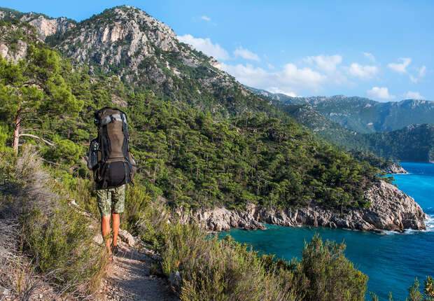A backpacker hikes along a narrow trail surrounded by lush greenery, overlooking a blue coastline and rugged mountains on a clear day.