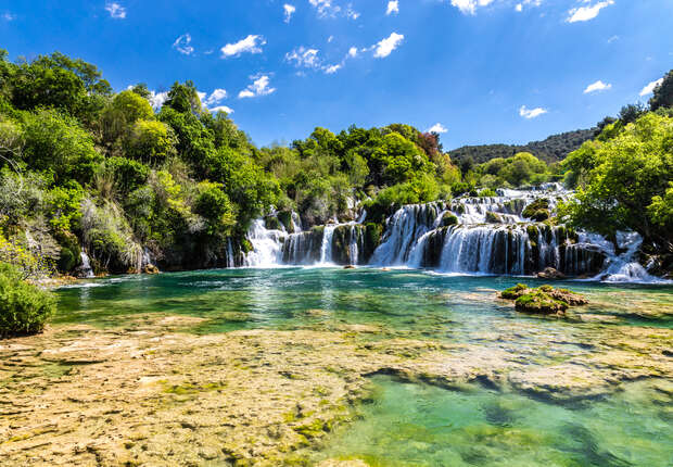 A multi-tiered waterfall cascades into a clear turquoise pool, surrounded by lush green foliage and hills under a bright, partly cloudy sky.