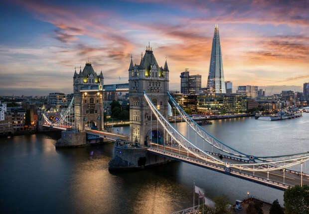 Tower Bridge, illuminated, spans a river during sunset. The Shard and city buildings form the urban backdrop, with a colorful sky enhancing the scene's dramatic ambiance.