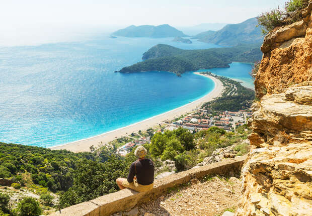A person sits on a stone ledge, overlooking a vast curved shoreline with turquoise water and green hills, while a small town nestles near the beach below.