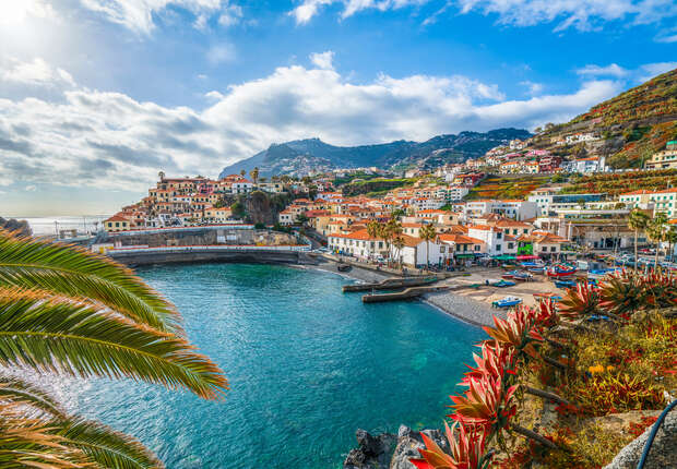 Coastal town overlooks calm bay with vibrant blue water; houses with red roofs cascade down hills. Lush greenery and colorful plants frame the scene under a partly cloudy, bright sky.