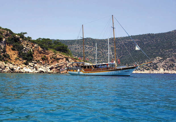 A wooden sailboat is anchored on tranquil blue waters, near a rocky coastline with sparse greenery. In the background, hills rise under a clear sky.