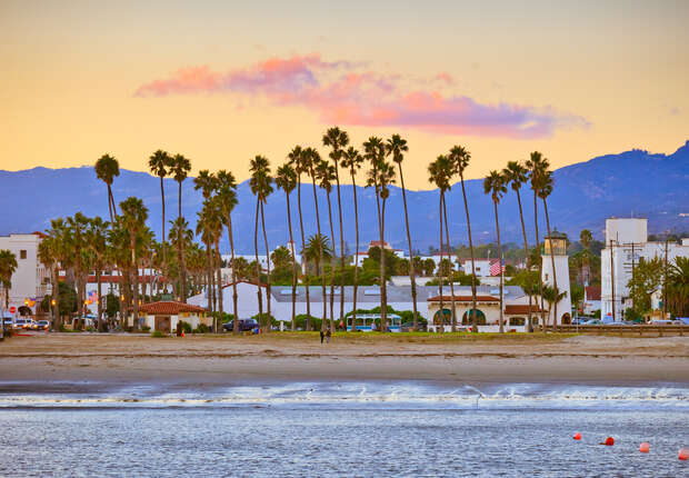 Tall palm trees stand along a sandy beach, with gentle waves in the foreground and a cityscape, including buildings and mountains, under a colorful sunset sky in the background.