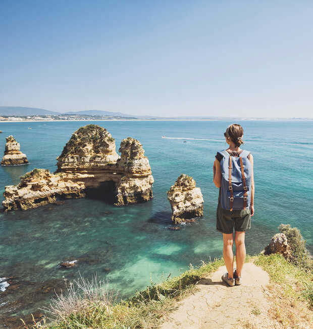 A person with a backpack stands on a cliff, overlooking rocky formations in turquoise ocean water, with a clear blue sky and distant coastline in the background.
