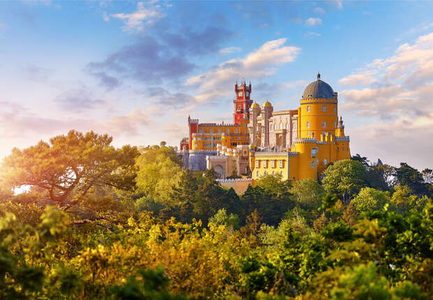 A colorful castle, featuring domes and towers, stands majestically atop a forested hill under a clear sky with scattered clouds.