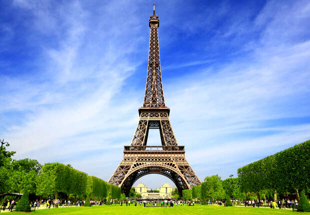 The Eiffel Tower stands tall against a blue sky, surrounded by lush green trees. People walk around its base in a scenic park setting.