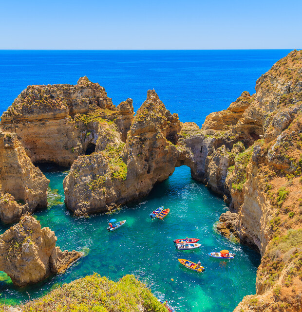 Kayaks and small boats navigate through a rocky coastal area with towering cliffs, surrounded by clear blue water and under a bright blue sky.