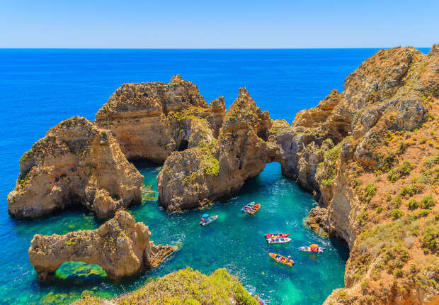 Boats float serenely in a crystal-clear turquoise sea, surrounded by rugged, towering cliffs and arch formations under a vibrant blue sky, creating a picturesque coastal scene.
