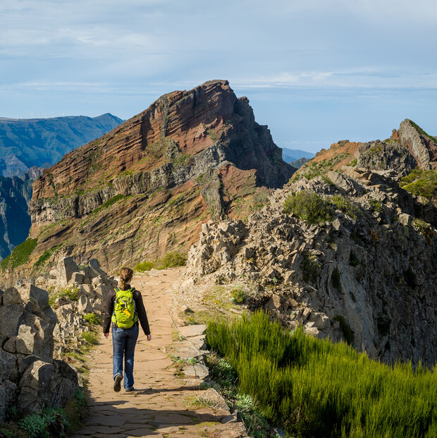 A person with a bright green backpack walks along a rocky mountain path, surrounded by rugged cliffs and vegetation under a clear sky.