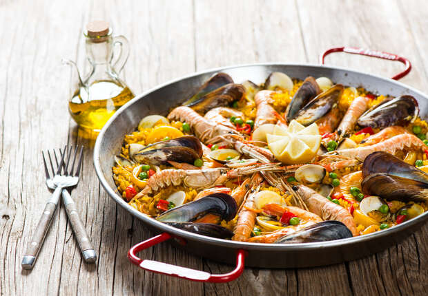 A large paella pan filled with seafood-topped rice is placed on a rustic wooden table. Two forks and a knife rest beside a small bottle of olive oil.