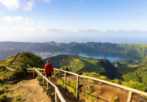 A person walks along a wooden fenced path overlooking green hills and a large blue lake, with distant mountains under a clear, sunny sky.