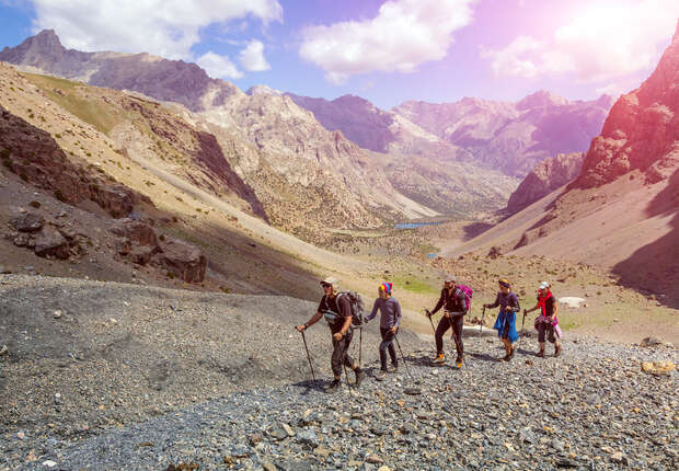 Hikers traverse a rocky mountain path, using trekking poles, surrounded by a vast valley with towering peaks under a partly cloudy sky. Sunlight highlights the scenic landscape.