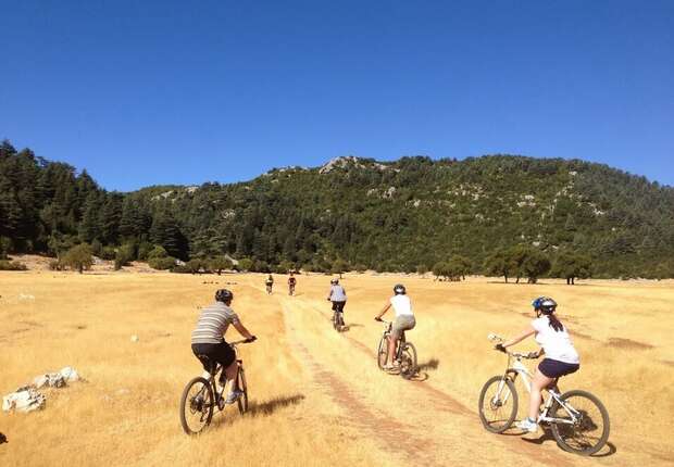 Cyclists ride along a dirt path in an open, grassy field surrounded by forested hills under a clear blue sky.
