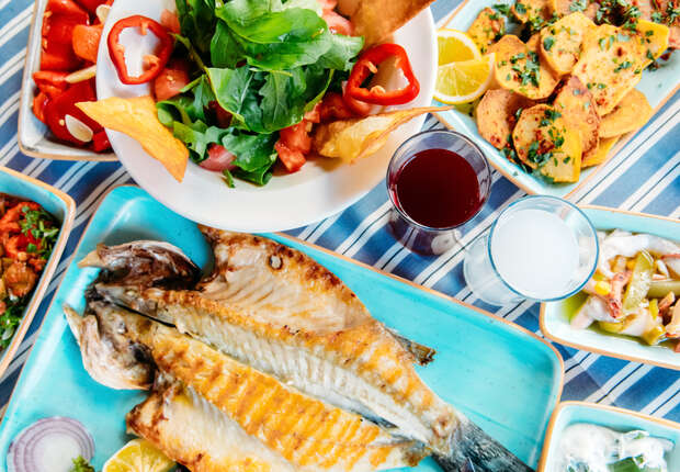 Grilled fish served on a blue plate, accompanied by a vibrant salad and seasoned potato slices, rests on a striped tablecloth alongside two glasses of red and clear beverages.