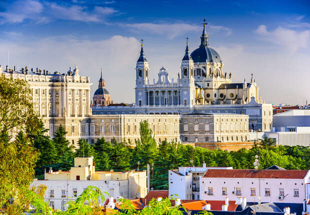 A grand cathedral with ornate domes and spires stands against a clear blue sky, surrounded by lush greenery and adjacent classical buildings, creating a serene urban landscape.