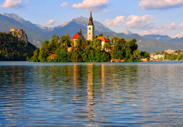 A church sits atop a small, tree-covered island, reflected in a calm lake, surrounded by distant mountains under a partly cloudy sky.