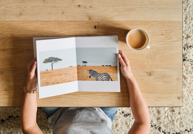 Hands hold an open book displaying a zebra in a savanna, with a tree visible. A coffee cup sits beside it on a wooden table, over a textured rug.