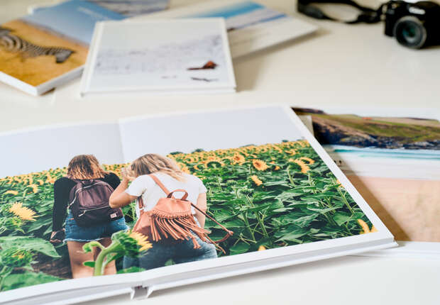 An open photo album displays two people walking through a sunflower field. Surrounding it, other albums with nature images lie on a light-colored table, along with a camera.
