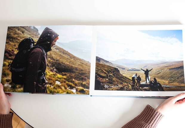 An open photo book on a table features a person hiking a grassy mountainous terrain on one page and a group of hikers celebrating atop a ridge on the opposite page.