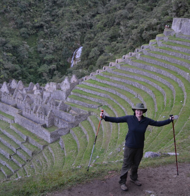 A person stands smiling with hiking poles on a dirt path before terraced ruins and lush forest, with a distant waterfall in the background.