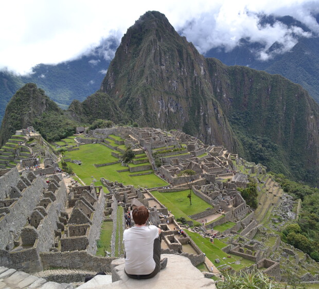 A person sits on a ledge, overlooking the ancient stone ruins of Machu Picchu, surrounded by lush green terraces and towering mountain peaks under a cloudy sky.