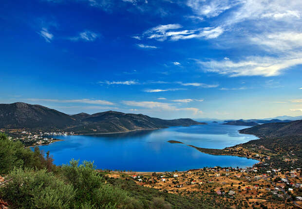 A serene blue lake reflects the sky, bordered by green hills. Small settlements and patches of vegetation dot the surrounding landscape under a vast, cloud-dotted sky.