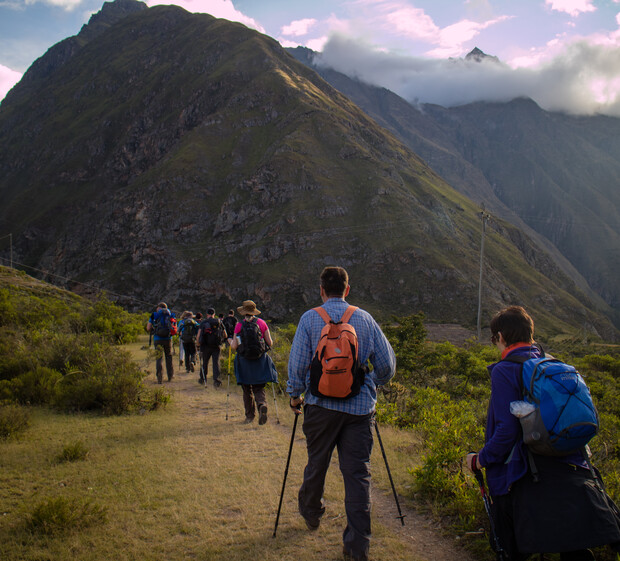 Hikers trek along a grassy path using poles, surrounded by lush greenery and dramatic mountain peaks under a partly cloudy sky.