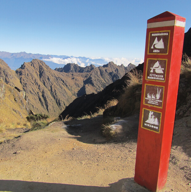 A red post stands displaying text: "WARMIWAÑUSKA ABRA 4215 m.s.n.m. WARMIWAÑUSKA PARQUE NACIONAL SANTUARIO HISTORICAL MIRADOR," against a backdrop of rugged mountain peaks under a clear blue sky.