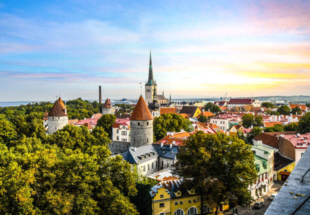 A picturesque medieval town features stone towers with red cone roofs amidst lush greenery, surrounded by colorful buildings under a vibrant sunset sky.