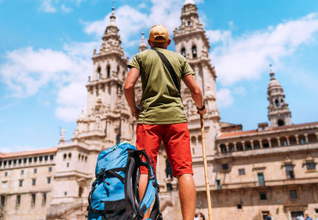A backpacker stands with a walking stick, gazing at a grand, historical cathedral on a sunny day, surrounded by ancient stone architecture.