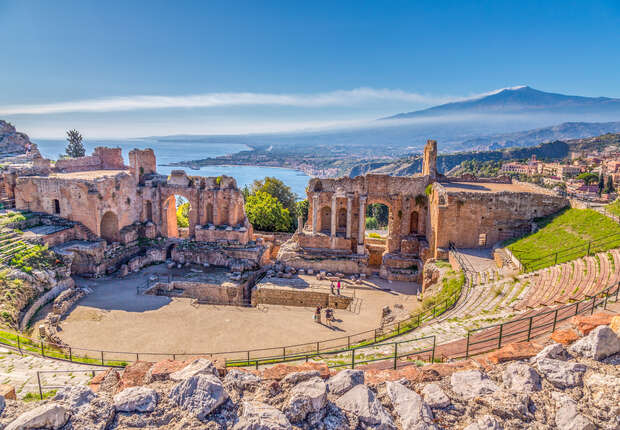 Ancient stone ruins stand overlooking a coastal landscape, with scattered visitors exploring the site. In the background, a distant mountain rises against a clear blue sky.
