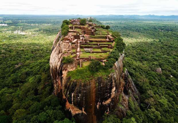 An ancient fortress and garden complex atop a massive rock formation, surrounded by dense green forest stretching towards distant hills under a cloudy sky.