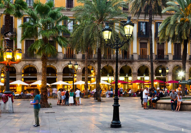People socialize near a central fountain in a lively plaza surrounded by palm trees and arched buildings. Lampposts illuminate the area as umbrellas shelter outdoor diners.