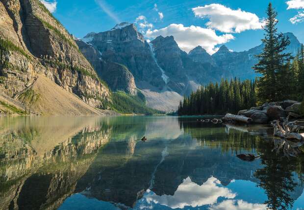 A calm lake reflects towering mountains under a blue sky with clouds, surrounded by dense pine forests. Logs rest near the shore, enhancing the tranquil, natural setting.