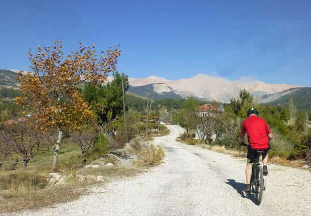 A cyclist wearing a red shirt rides along a gravel path through a scenic rural landscape with trees and distant mountains under a clear blue sky.