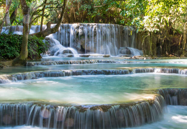 Cascading waterfalls flow gracefully over tiered rock formations, surrounded by lush green foliage and dappled sunlight, creating a serene, natural scene in a forested area.