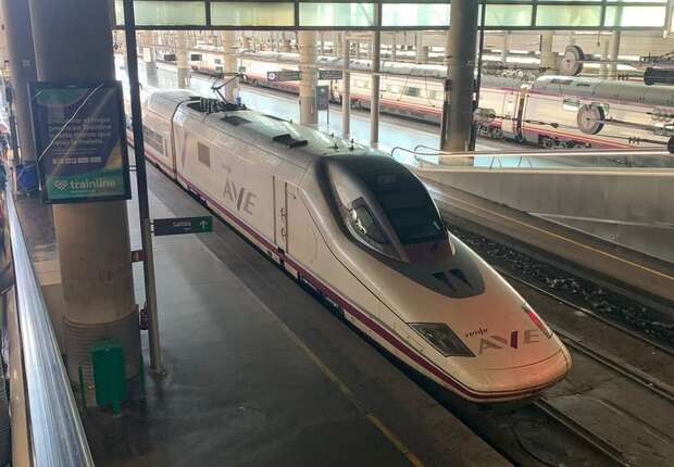 A high-speed AVE train is stationary on a platform in a spacious, modern train station. Signs nearby display "trainline" and an arrow with "salida" on a green background.