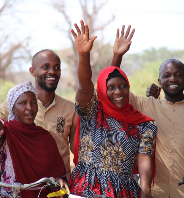 Four people stand outdoors, smiling and waving. The two women in front are wearing headscarves, one red and one patterned. Two men in beige shirts stand behind them. Trees are in the background.