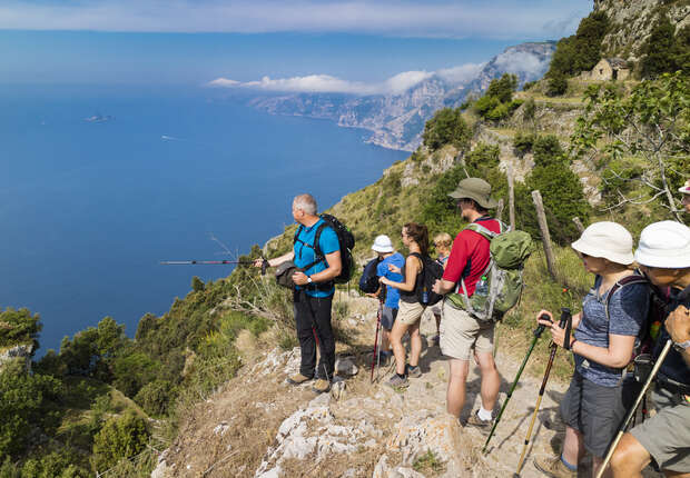 Hikers pause on a narrow, rocky trail along a steep cliff, overlooking a vast blue sea and distant islands, surrounded by lush greenery and mountains under a clear sky.