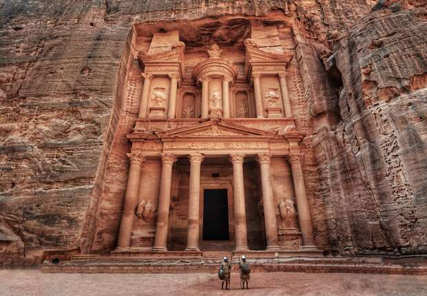 A grand ancient stone building with intricate carvings and columns is nestled in a rocky cliff face; two people on camels stand in front, highlighting its scale and historical context.