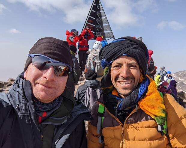 Two people in winter gear pose atop a snowy mountain, smiling. Behind them, a group of climbers gathers around a metal pyramid structure under a clear, cloudy sky.