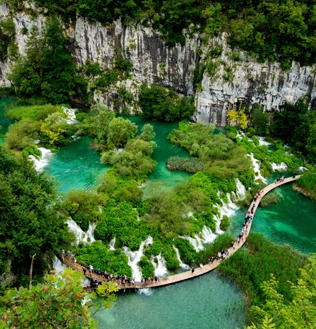 A curved wooden walkway crosses vibrant turquoise lakes with cascading waterfalls, surrounded by lush, green foliage and rocky cliffs; people stroll along the path, admiring the scenic landscape.