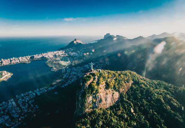 A large statue stands atop a lush, green mountain overlooking a sprawling city by the ocean, with hills in the background under a blue sky.