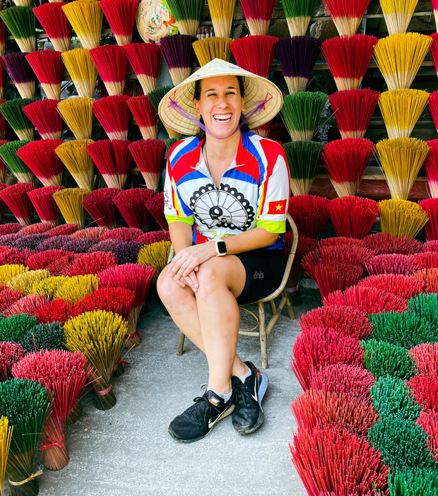 A person wearing a conical hat and colorful shirt, sitting on a stool, smiles surrounded by vibrant bundles of incense sticks displayed in various colors against a wall.