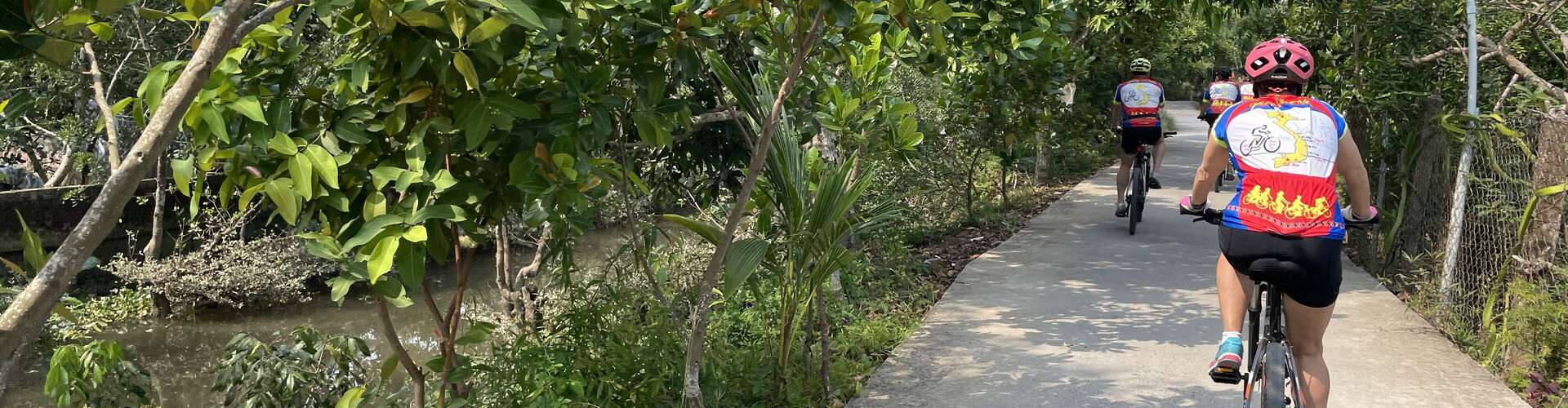 Cyclists ride along a narrow, tree-lined path beside a small river, wearing brightly colored jerseys with a bicycle graphic and map design, amidst lush green foliage.