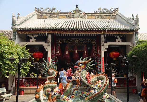 An ornate dragon sculpture stands in a courtyard fountain, surrounded by visitors outside a traditional Chinese building with red lanterns and intricate carvings, under a clear sky.