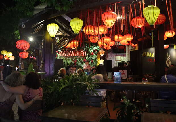 Colorful lanterns hang decoratively, illuminating a lively, bustling restaurant at night. People sit, eat, and socialize under the warm glow. Prominent signs read "MADAM KIEU," adding an inviting ambiance amid lush greenery.